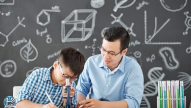 طالب صغير يستخدم الميكروسكوب بمساعدة معلم أمام سبورة مملوءة برموز فيزيائية وكيميائية، في بيئة تعليمية محفزة.