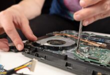 شخص يقوم بصيانة جهاز كمبيوتر محمول مفتوح باستخدام مفك، ويظهر مروحة التبريد واللوحة الأم.