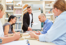 أشخاص يجلسون حول طاولة في مكتبة ويلعبون ألعاباً جماعية بالورقة والقلم ضمن أجواء تفاعلية ممتعة.