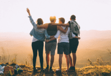 أصدقاء يقفون على قمة جبل عند الغروب يحيطون بعضهم بالأذرع في مشهد يعبر عن الصداقة الحقيقية.
