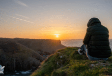 شخص يجلس على حافة منحدر يطل على البحر أثناء غروب الشمس، في لحظة تأمل وهدوء.