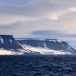 مشهد طبيعي من سواحل المحيط المتجمد الشمالي يظهر فيه الجليد والجبال المغطاة بالثلوج تحت سماء غائمة.